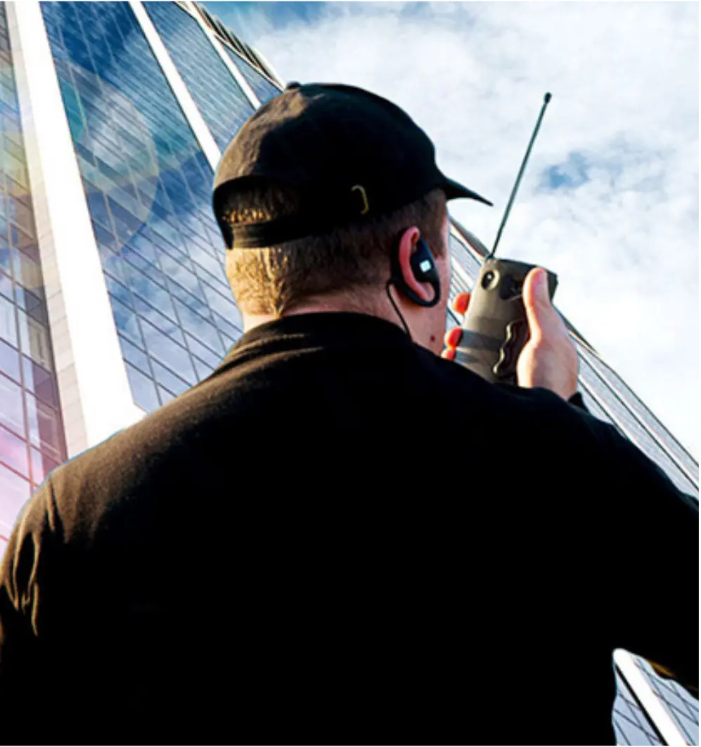 Pessoa de costas usando um boné preto e fones de ouvido, segurando um walkie-talkie, com um prédio alto de fundo